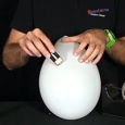 Someone's hands wearing a green wrist watch, holding a 9 volt battery that is held up to a inflated white balloon.