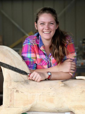 A young woman named Olivia leaning on a unpainted rocking horse holding a wood filing tool.