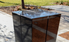 A rectangular black granite pilar about 1500 milimeters heigh, with for slits running from across the top to three quarters of the way down the sides. The pilar sits on concrete and is located in a garden environment.