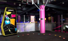 A multi coloured sign with the words 'Optimusic' at the top, along side a carpeted area with a pink structural pole, and different coloured floor lights in a curved line.