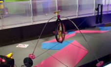 A wheel sits on a cable tightrope, with two masses extending from the wheel so they hang below the cable tightrope