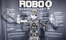 A robot with it's arms spread outwards, and a large grey and white information panel behind it with the title 'Robo Q Robothespian'.