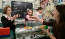 Children can observe, imitate and invent make-believe situations (which develops imagination and creativity). Three young girls playing in a toy bakery, and serving a woman.