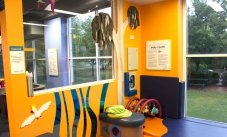 A special space for babies and their carers. A brightly coloured room of yellow, blue and purple, with windows looking to outside garens, and soft play matts and baby toys on the ground.