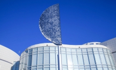 A large D shaped silver sculpture sitting on a pole with thousands of small metal round discs attached. Behind is a curved white building with many windows and a blue sky.