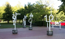 Four white steel outline scupltures of people on a paved area with gardens in the background.