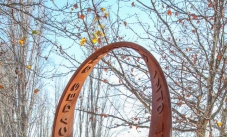 Select one spot on the sculpture and imagine tracing a toy car running around the track and returning to the starting position. A rusted continuous steel loop sculpture with the raised words 'looping cascades' and 'into sprial worlds' on the surface of the strip. The sculpture sits on a brown graveled garden area, with deciduous trees in the background.