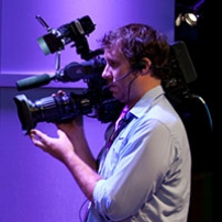 a man in front of a purple wall, holding a professional video camera.