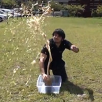 A man kneeling on a lawn, with a plastic tub and soft drink bottle in front of him. Soft drinking is projecting into the air.