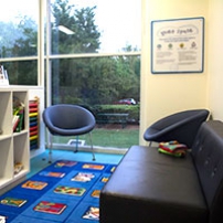 A light lit room with a bookshelf and books, a play rug, a leather couch, two seats and a window looking to the outside garden.