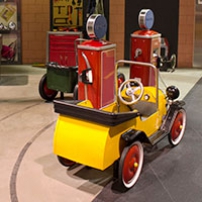 A yellow ride on toy car with red wheels parked along side replica toy red petrol bowsers.
