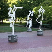 Three white steel outline scupltures of people on a paved area with gardens in the background.