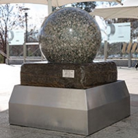 A large granite stone ball sitting on a large slab of stone, which intern sits within a stainless steel bin. Water flows from under the granite and over the stone to the bin.