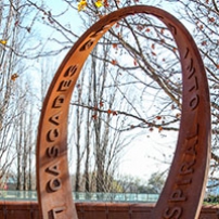 A rusted continuous steel loop sculpture with the raised words 'looping cascades'  and 'into sprial worlds' on the surface of the strip. The sculpture sits on a brown graveled garden area, with deciduous trees in the background.