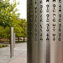 Three stainless steel vertical poles about 2 metres in height are spaced apart and embedded into a concrete and paved garden area. On each of the poles are laser cut out cryptographic symbols.