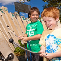 Two young boys each with a metal mallet and rubber mallet are hitting semi-vertical stones notes arranged in a keyboard arrangement.
