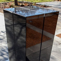 A rectangular black granite pilar about 1500 milimeters heigh, with for slits running from across the top to three quarters of the way down the sides. The pilar sits on concrete and is located in a garden environment.