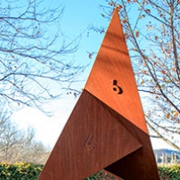A rusted metal sculpture with six faces located in a garden environment.