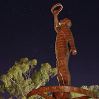 A night time photo of a rusted metal sculpture of a man holding a ring up to the sky with one outstretched arm. There are gum trees lit up in the background and the sky is black with thousands of stars visible.