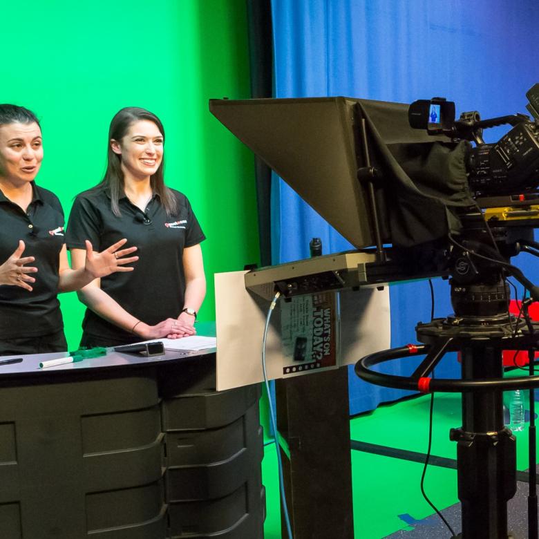 Two presenters, wearing black Questacon branded shirts, stand in front of a large green screen. One of the presenters are talking into a large studio video camera.