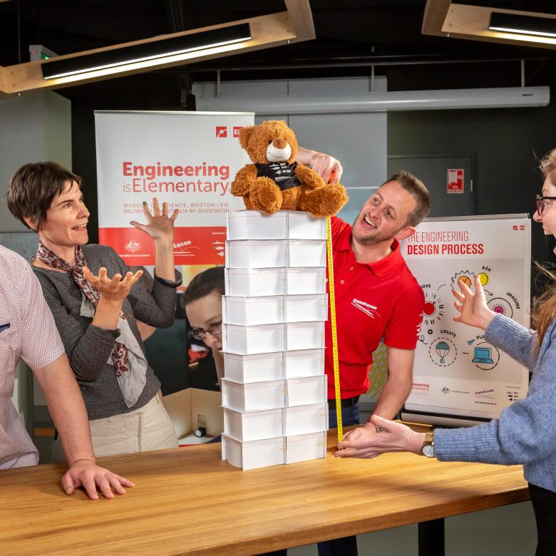 3 teachers have built a tower using cards of paper to balance a bear on it. The Engineering is Elementary facilitator is measuring their work. All are displaying emotions of happiness as the tower stays up.