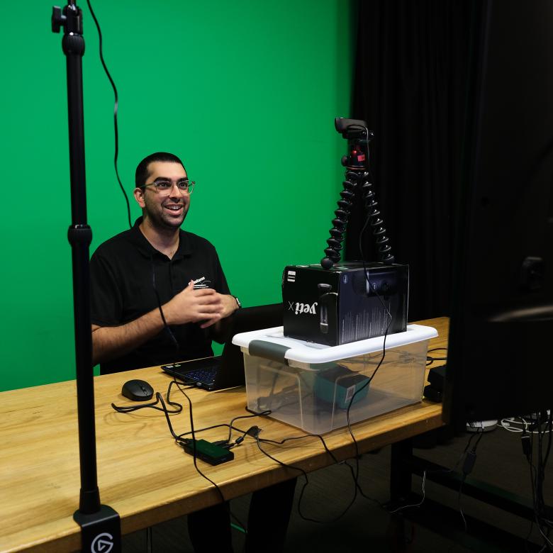 Questacon presenter in front of a green screen, smiling towards a filming equipment.