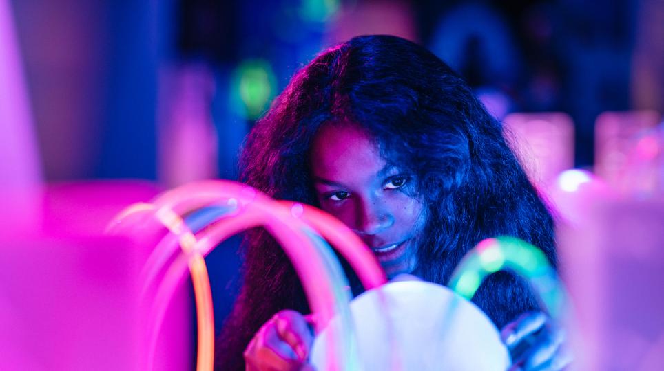 A person sits at a glowing interactive table surrounded by neon-coloured light tubes, focusing on an illuminated object in front of them.