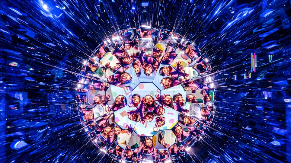 A large kaleidoscope-style installation forms a spherical pattern of repeating reflections of people reaching toward its centre, surrounded by streaks of bright blue light.