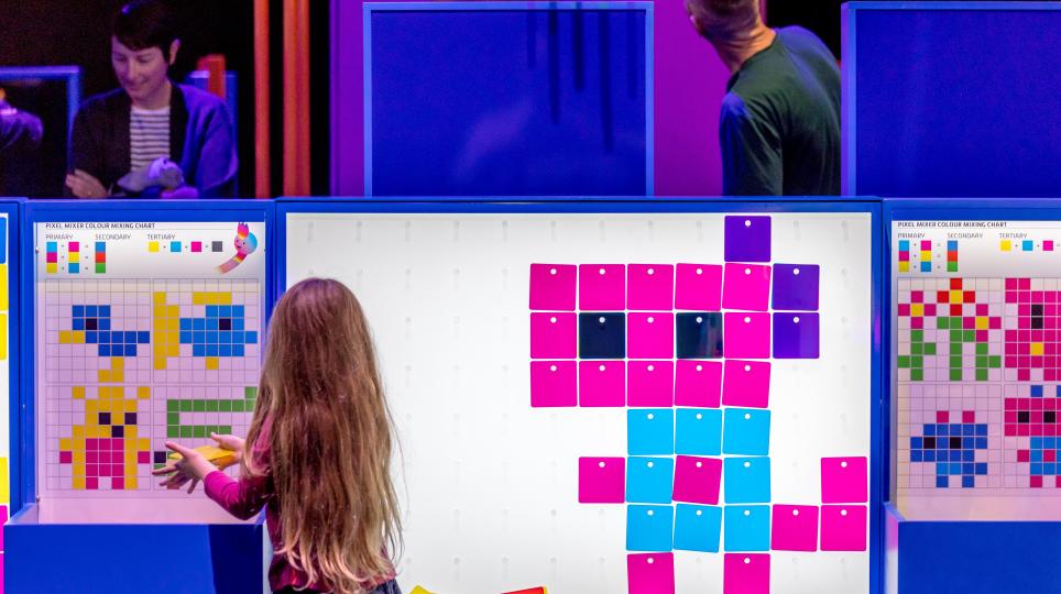 A child stands at an illuminated panel creating pixel‑style artwork using brightly coloured square tiles. Additional mosaic-style creations are displayed on panels nearby.