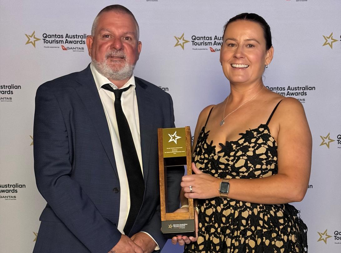 Two people standing in front of a Qantas Australian Tourism Awards 2025 media wall, dressed in formal attire. One person is holding a tall gold-and-black trophy featuring a star emblem and award text. Both individuals are posed for an official event photo.
